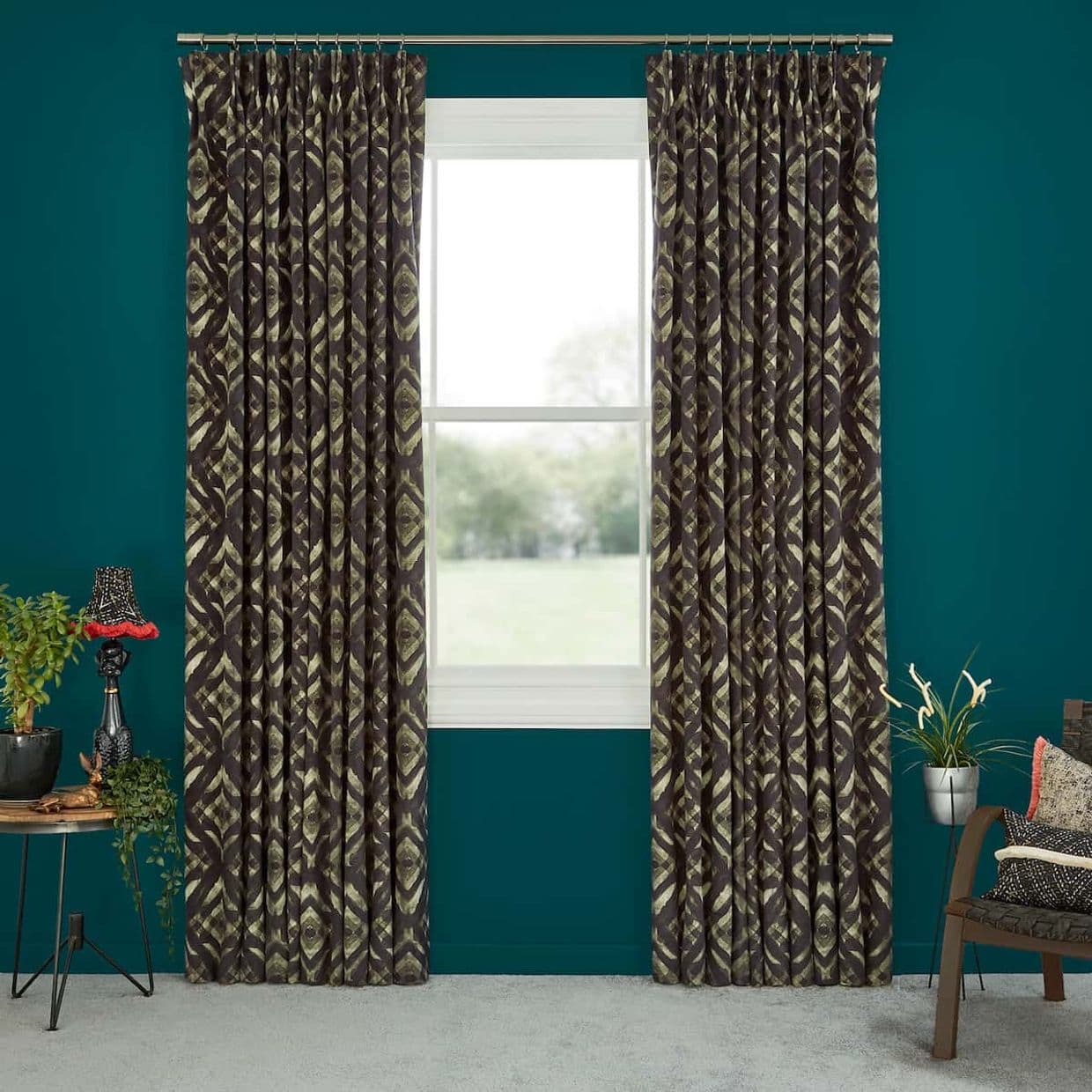 Pleated dark green-and-olive ikat curtains on a metal rod, drawn open to frame a bright outdoor view; teal-painted room with plants, a lamp, and a chair.