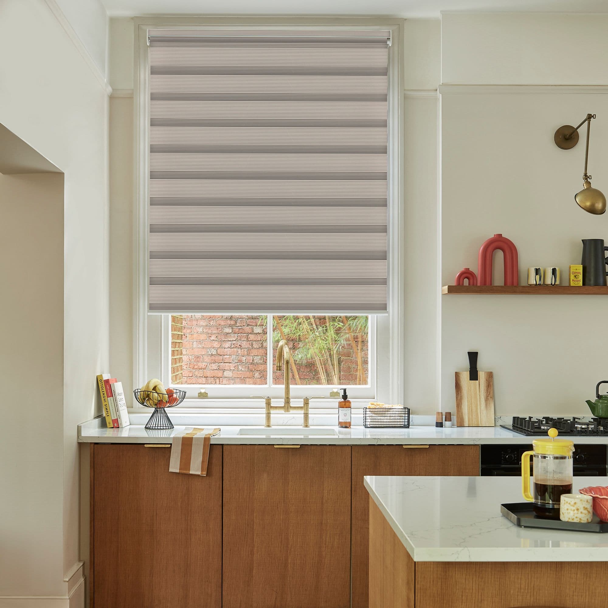 Beige pleated roller blind lowered across the window, gently filtering daylight; kitchen sink with brass faucet, marble countertops, wooden cabinets, shelf decor, and a small brick-and-plant courtyard view.