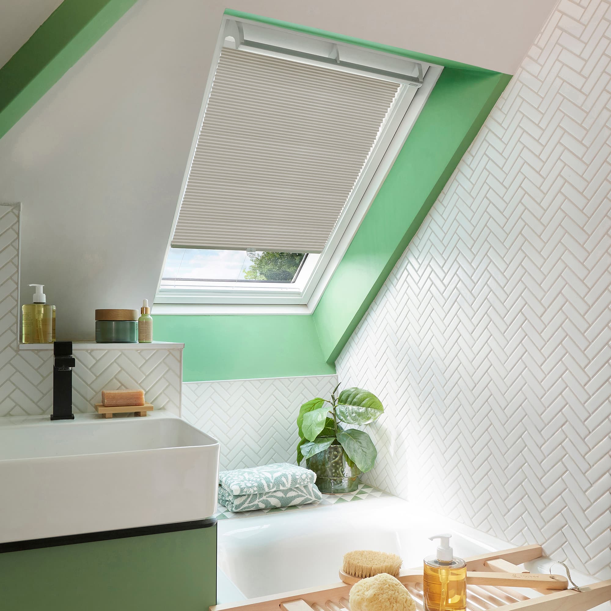 Pleated white skylight blind, partially lowered over a slanted roof window; filtering soft daylight into a green-accented bathroom with white herringbone tiles, sink, bathtub, plant, and bath tray.