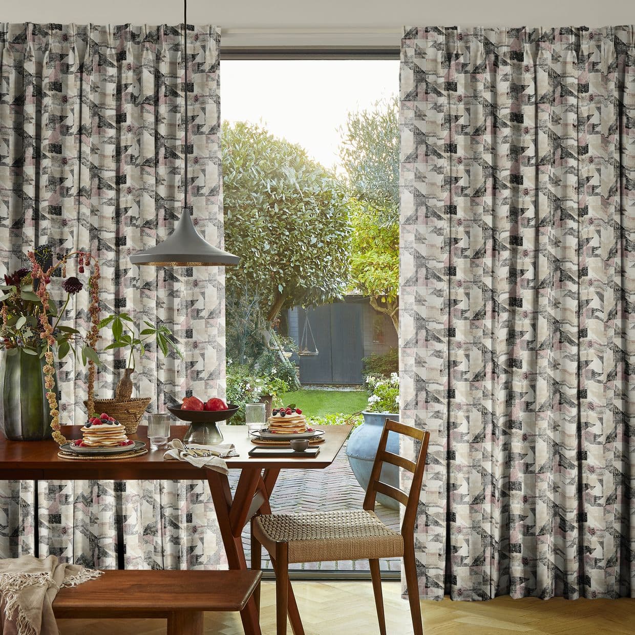 Pleated geometric-patterned curtains, drawn to either side, framing a central glass door; they partially filter light while revealing a sunlit backyard, with a dining table set for breakfast in the foreground.