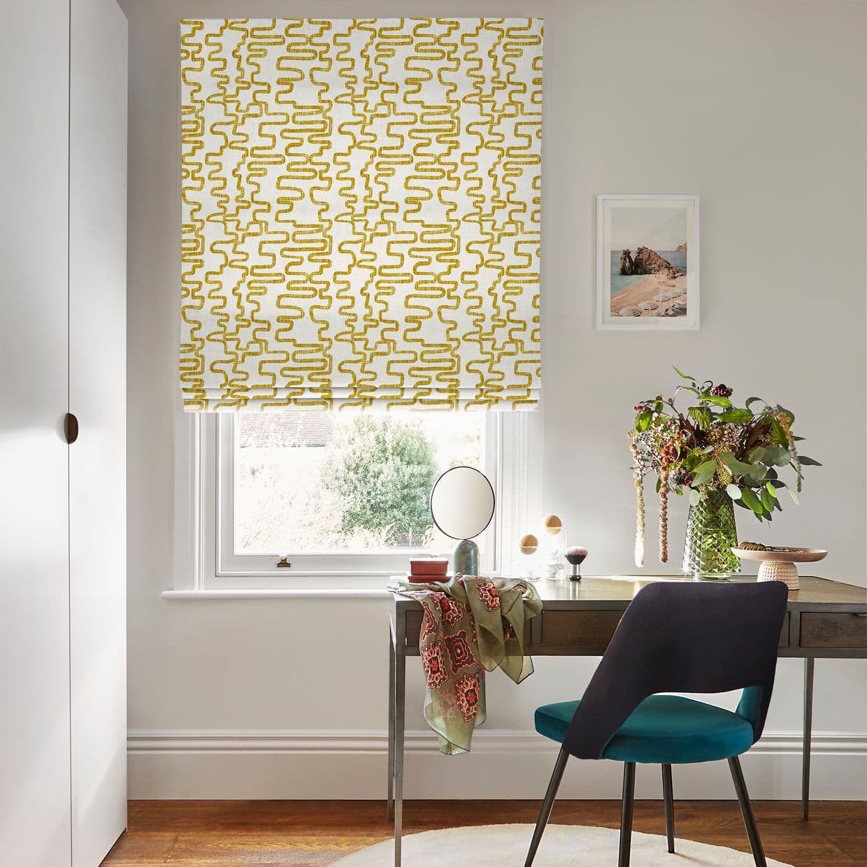 A patterned Roman blind (white with mustard-yellow abstract squiggles) is lowered over a sash window, filtering daylight. Context: pale room with wooden desk, teal chair, vase of foliage, mirror, framed coastal print.