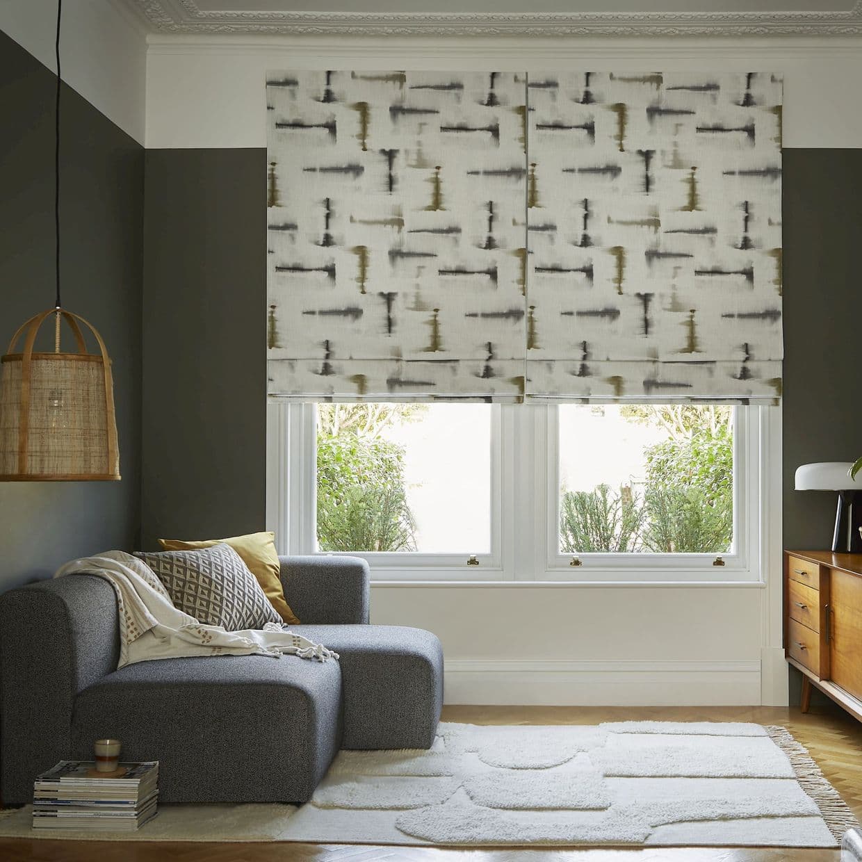 Patterned fabric Roman blindsâ€”paired over twin windows with abstract grayâ€‘olive brushstroke printâ€”are partially lowered, softly diffusing daylight; cozy living room with gray chaise, woven pendant lamp, wooden sideboard, garden view.