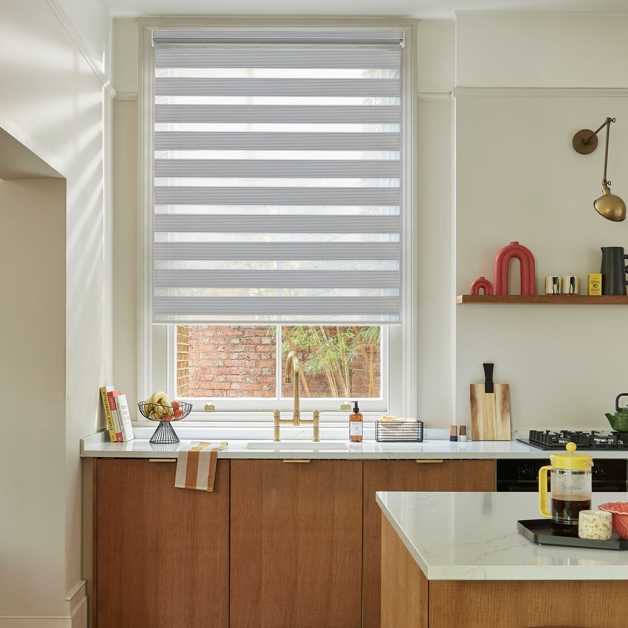 Horizontally striped dual-layer roller blind, lowered and filtering light; over a kitchen sink with brass faucet, wood cabinets, marble counters, an island, and a small brick courtyard visible outside.