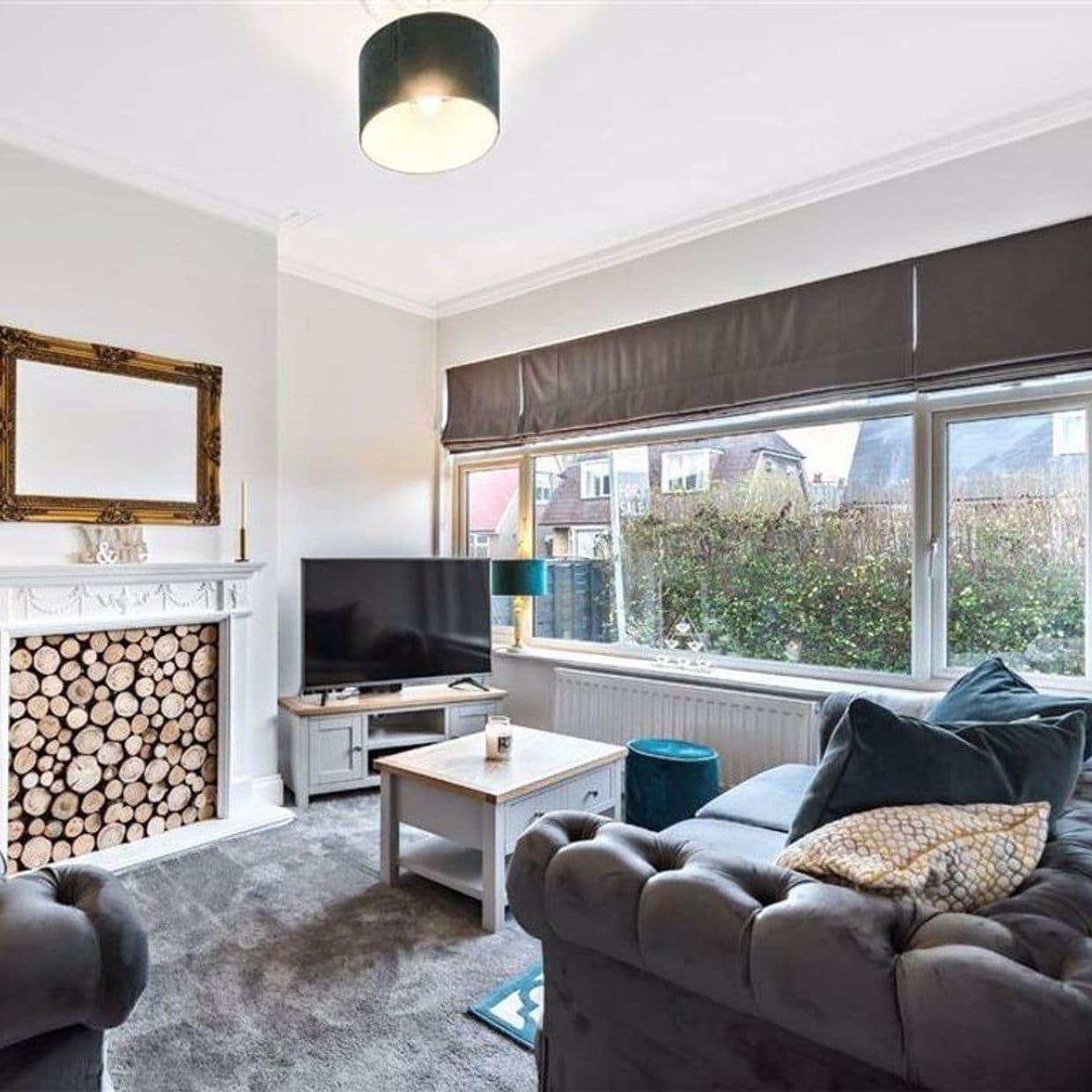 Dark gray Roman blind lowered across a wide front window, partially filtering daylight; living room with tufted gray sofa, TV, coffee table and decorative stacked logs in the fireplace, hedge and houses outside visible.

FOR SALE