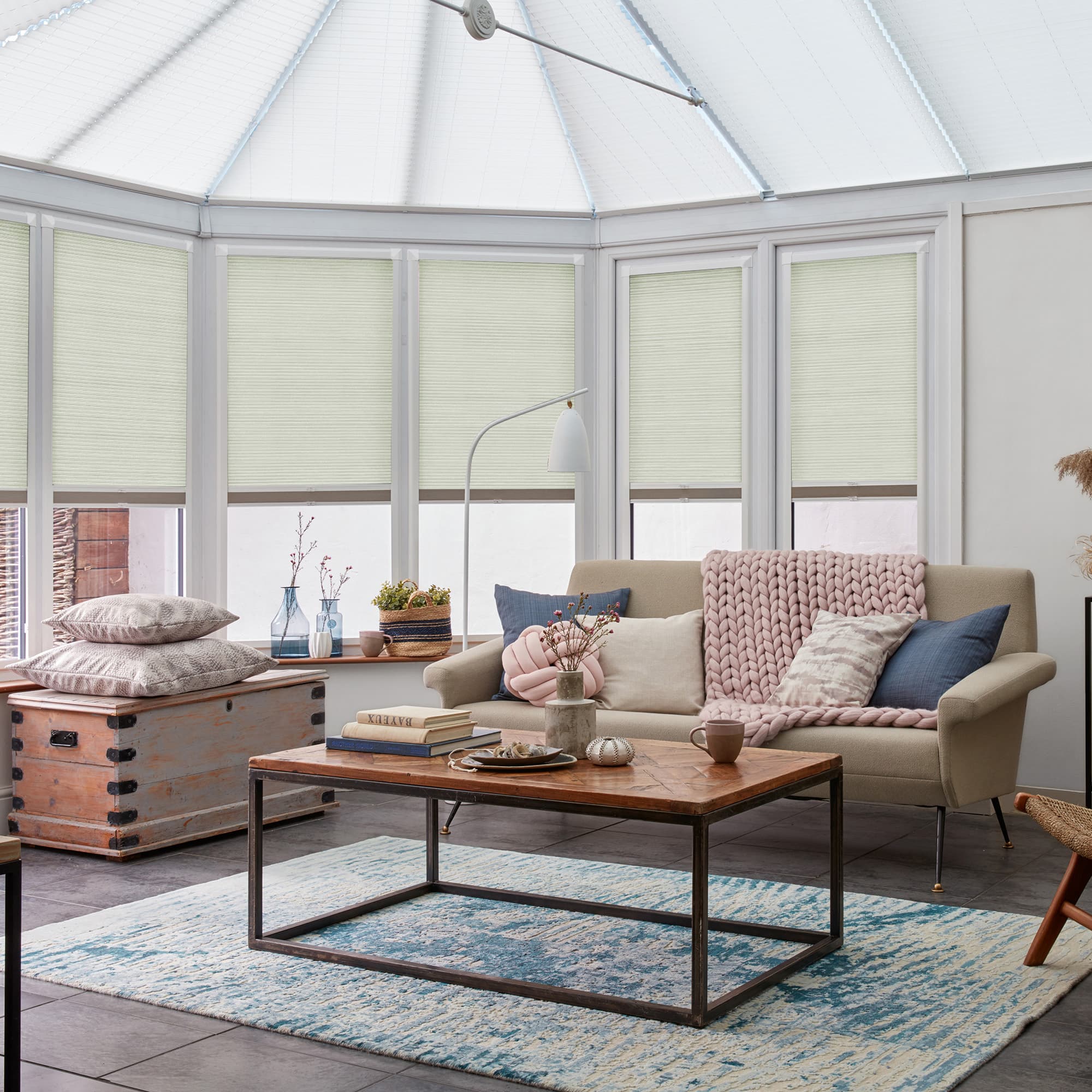 Light-colored pleated cellular blinds, lowered across angled conservatory windows, diffusing bright daylight. Cozy living area beyond: beige sofa with chunky pink throw, wooden coffee table and blue patterned rug.