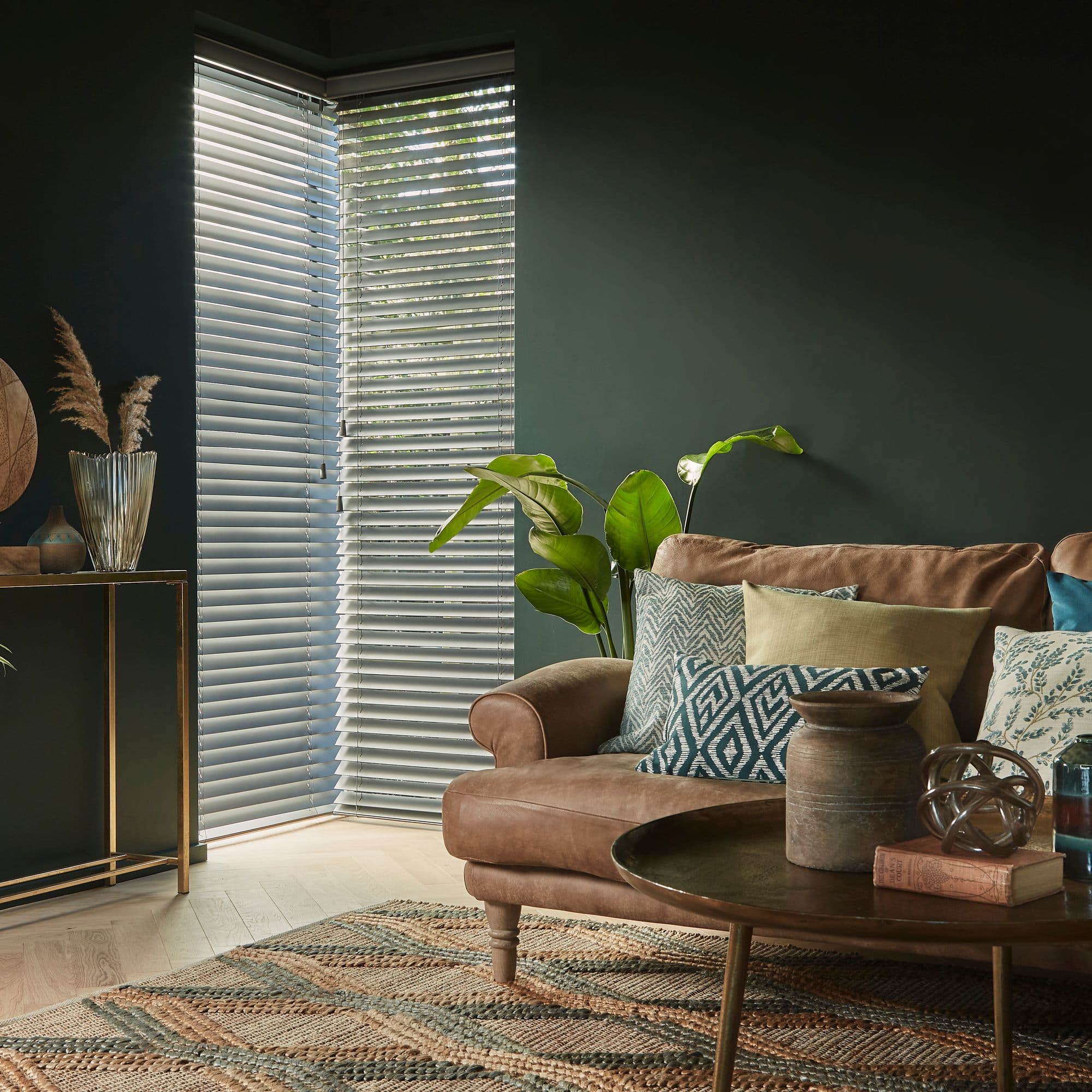 White horizontal blinds on a corner window, slatted and partially closed, filtering soft daylight into a dark-green living room with a brown leather sofa, patterned cushions and a coffee table.