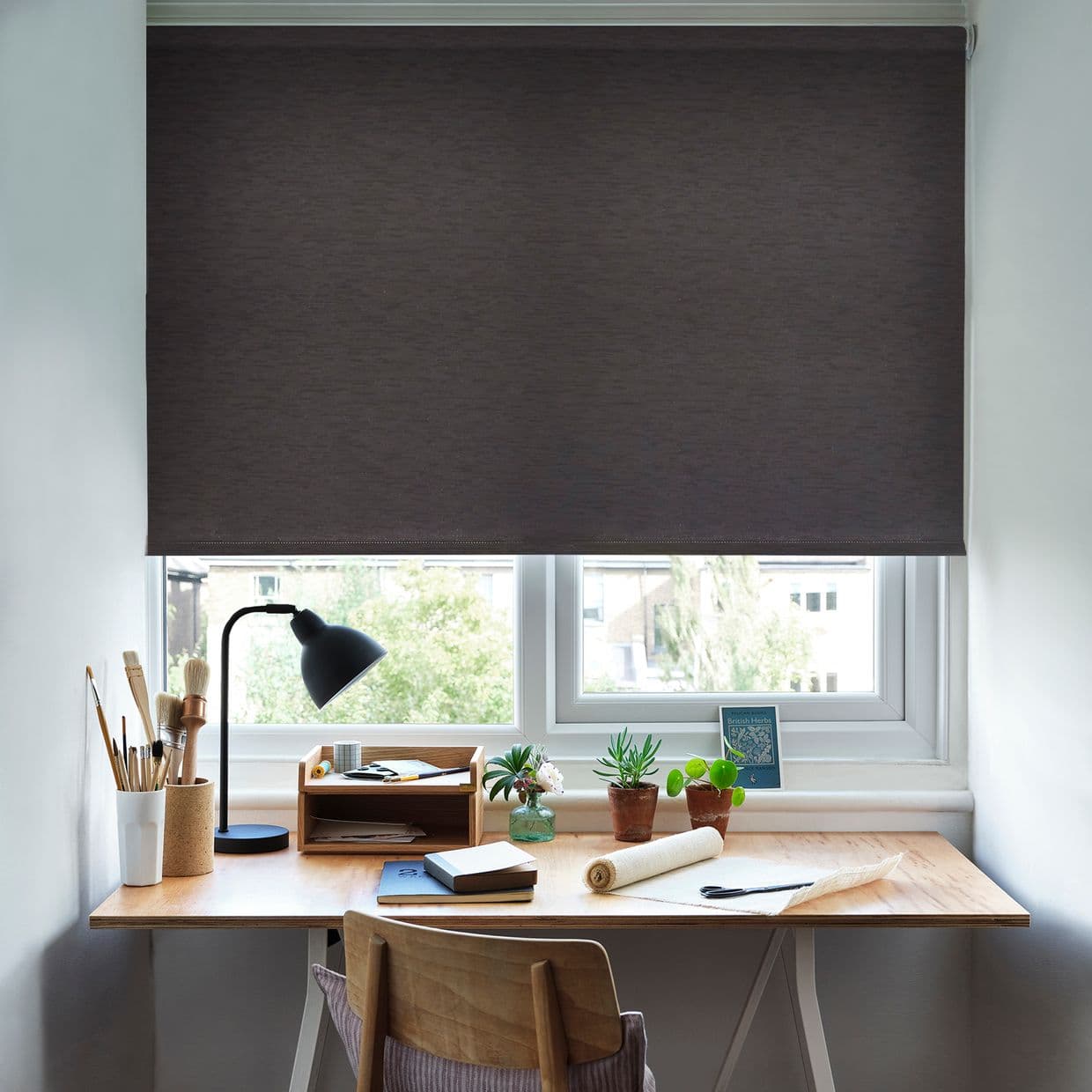 Dark grey roller blind lowered over a window, mostly closed and blocking light; below, a wooden desk with lamp, brushes, notebooks and potted plants in a bright home study.