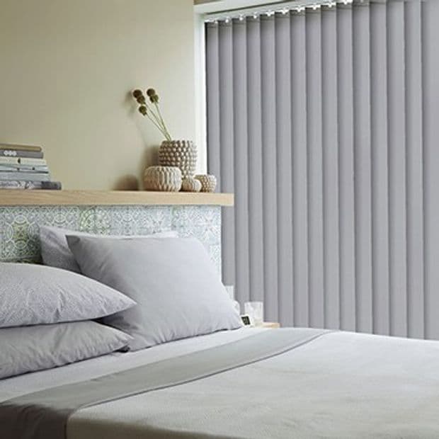 Vertical fabric blinds covering a full-height window at right, closed and filtering daylight; bedroom setting with neatly made bed, pale grey bedding, decorative shelf and small vases.