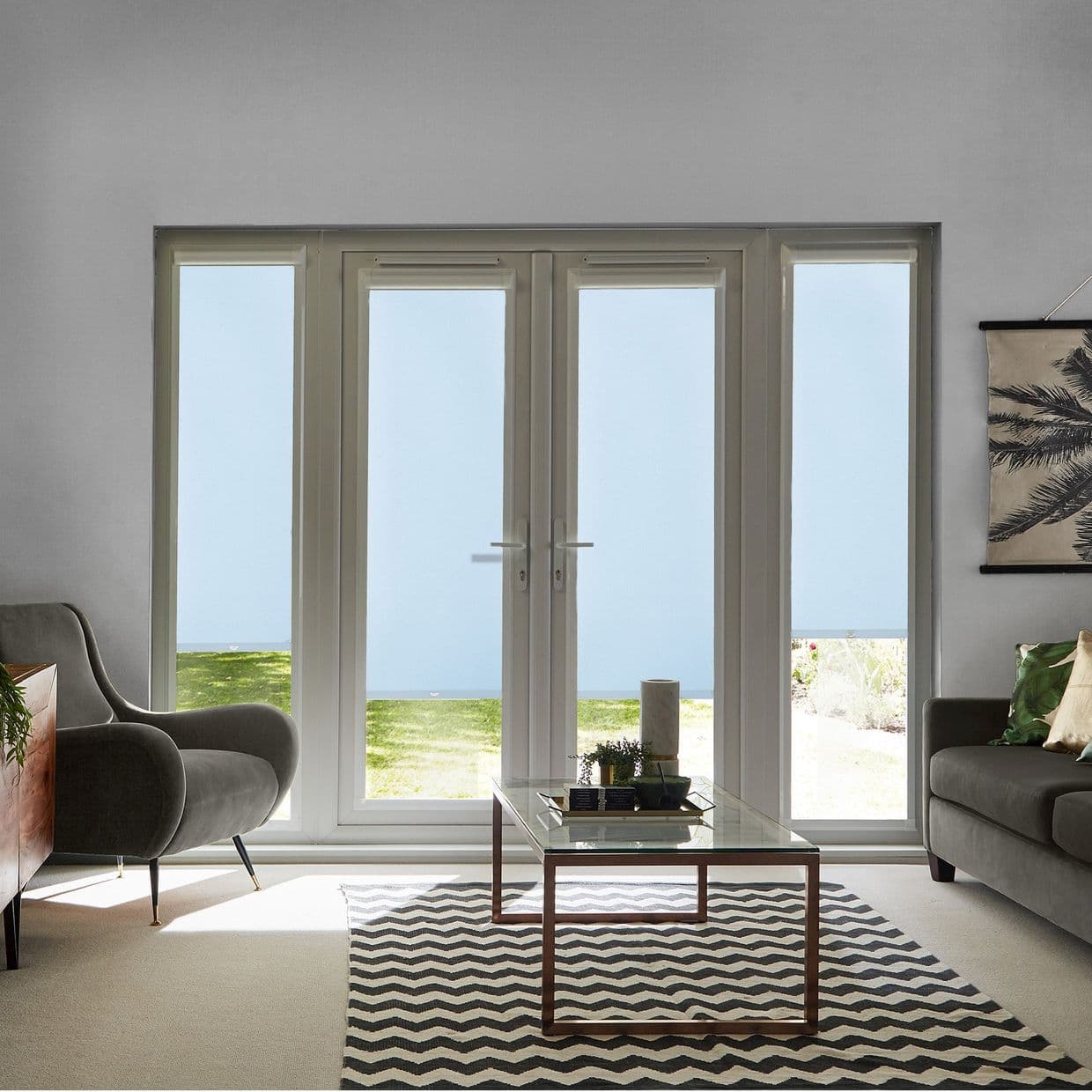 White-framed double French doors with narrow sidelights, closed and admitting diffused daylight; reveal lawn and distant water. Context: bright living room with armchairs, glass coffee table on chevron rug.