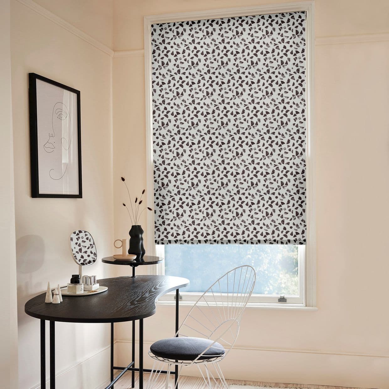 Patterned roller blind (white with scattered black terrazzo-like shapes) fully lowered, filtering daylight into a pale, minimalist room with a small black desk, wire chair, and framed line-art.