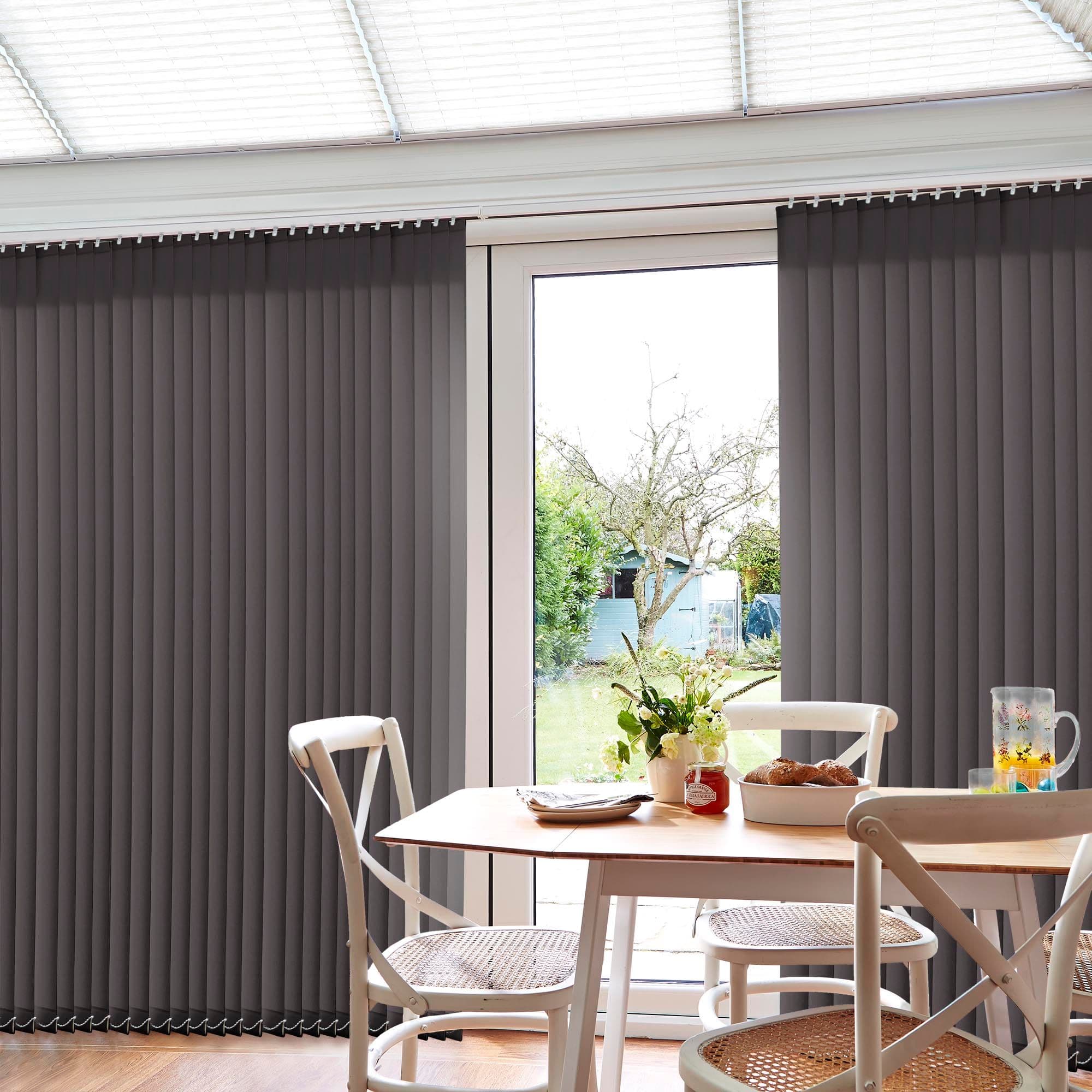 Grey vertical blinds covering a patio door, mostly closed with a central gap revealing glass; filtering daylight into a dining area with wooden table, chairs, vase, garden beyond.