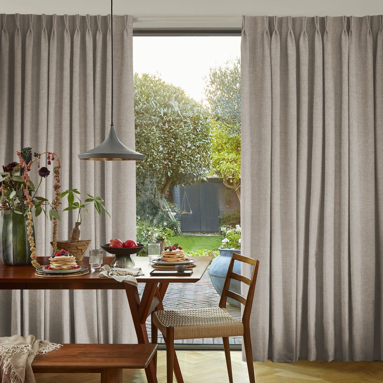 Full-length beige pleated curtains parted at center, framing a glass door and letting daylight through; wood dining table set with pancakes and pendant lamp overlooks a leafy garden.
