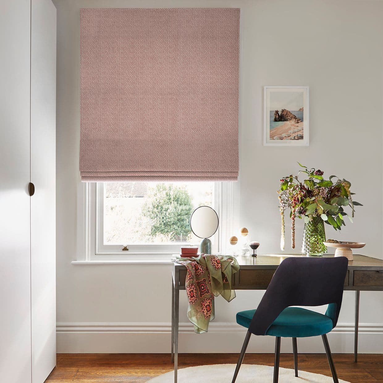 Pink textured Roman blind lowered over sash window, gently filtering daylight; light home office with wooden desk, teal chair, round mirror, vase of flowers, and garden visible beyond.
