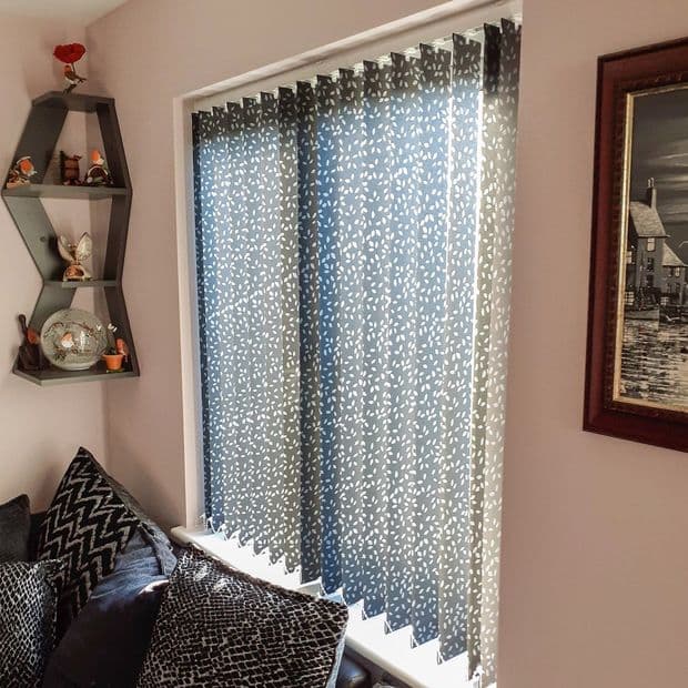 Patterned blue-and-white vertical blinds covering a window, closed and filtering daylight; set in a living-room corner above a sofa with patterned cushions, decorative hexagonal shelf and framed wall art.