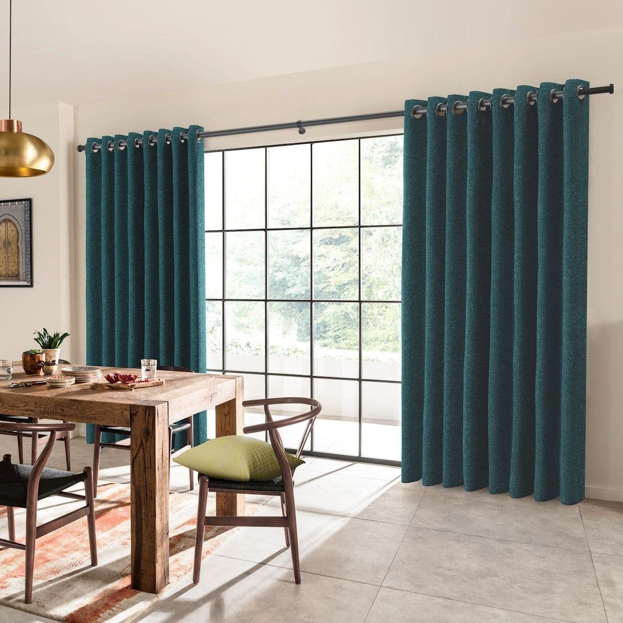 Teal grommet-top curtains, hung on a black metal rod, pulled to sides and framing a large grid-pane window; bright daylight floods a dining room with wooden table, chairs, and a pendant light.