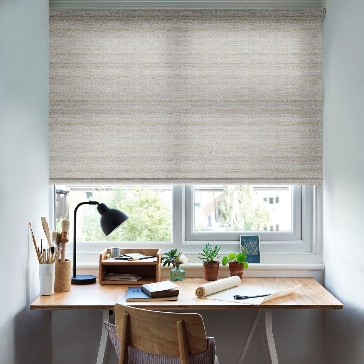 Beige textured roller blind lowered across a double window, diffusing daylight; a wooden desk beneath holds a black lamp, brushes, notebooks and small potted plants in a bright workspace.