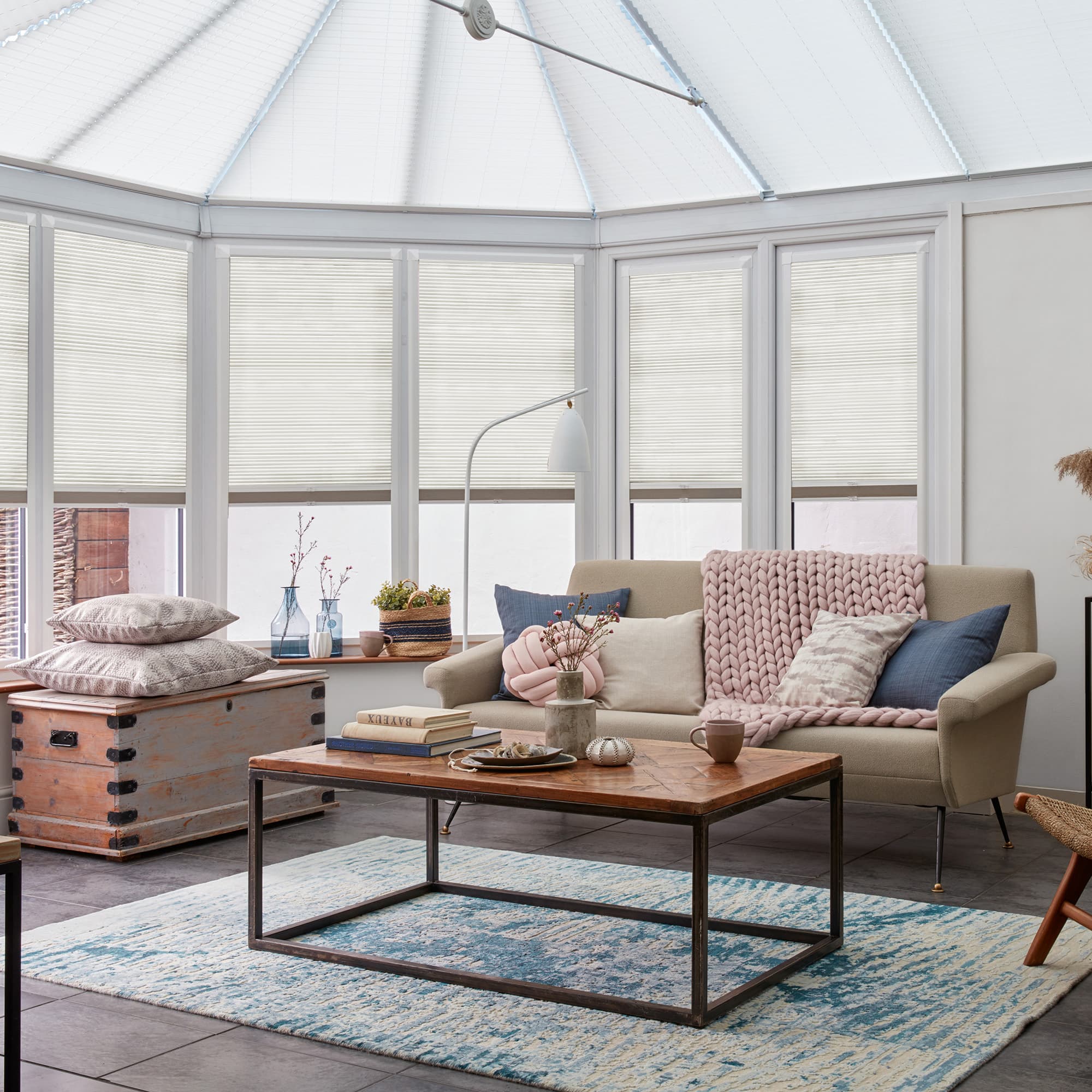 White pleated blinds on bay windows and roof, lowered and diffusing daylight; in a bright conservatory-style sitting area with neutral sofa, pink throw, wooden coffee table, blue rug.