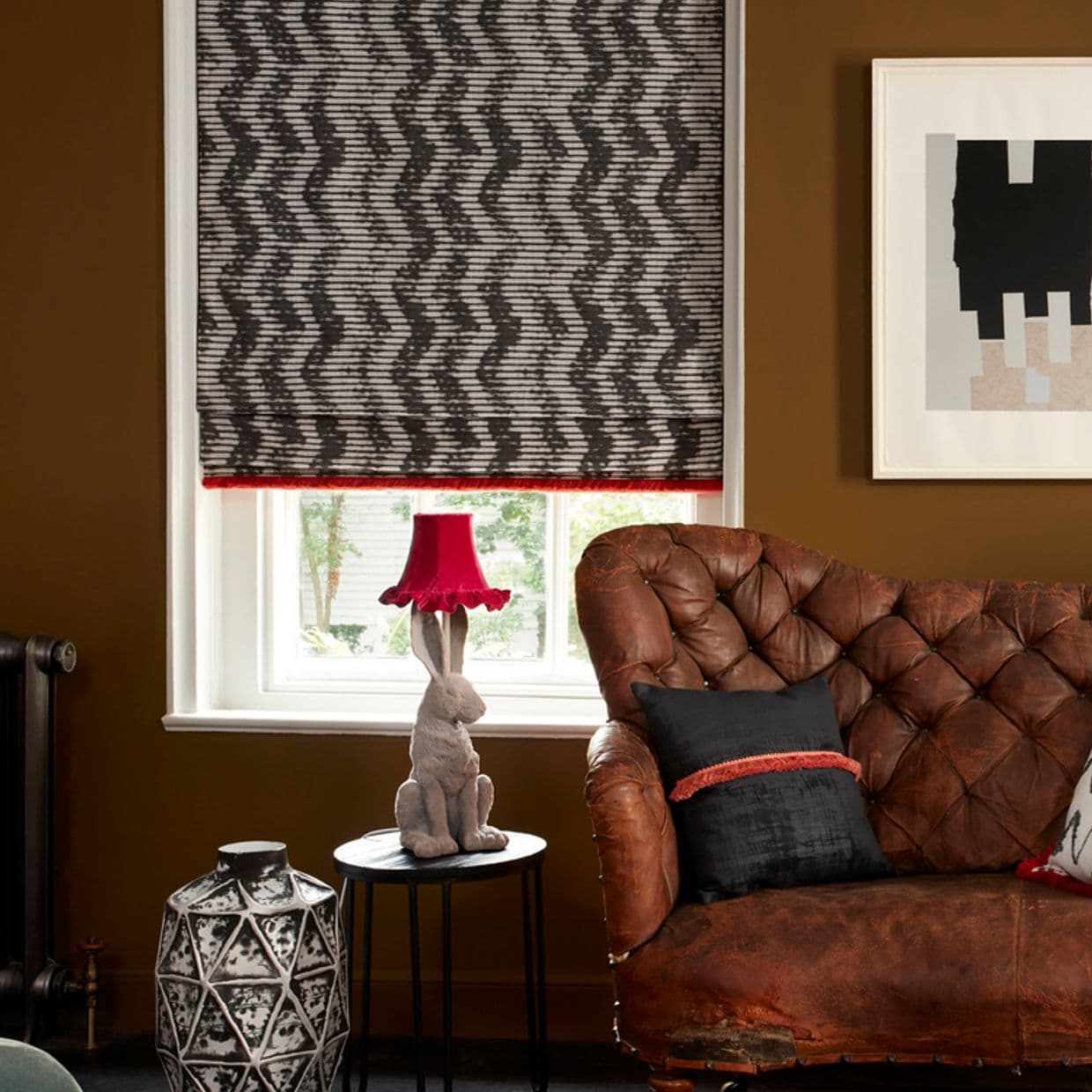 A patterned fabric Roman blind, lowered with black-and-white zigzag print and red trim, partially filtering daylight; set above a window in a warm-toned living room with a tufted leather sofa.