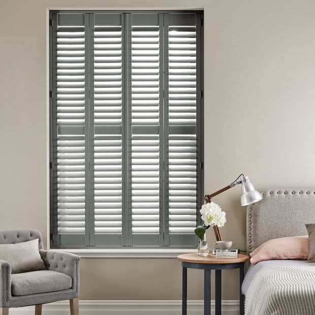 Gray plantation shutters covering a recessed tall window, four louvered panels closed with slats angled to filter daylight; set in a beige bedroom with armchair, bedside table, lamp, and flowers.