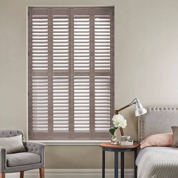 Grey wooden plantation shutters cover a tall window in four louvered panels; closed with slats angled to filter soft daylight into a neutral bedroom with armchair and bedside lamp.