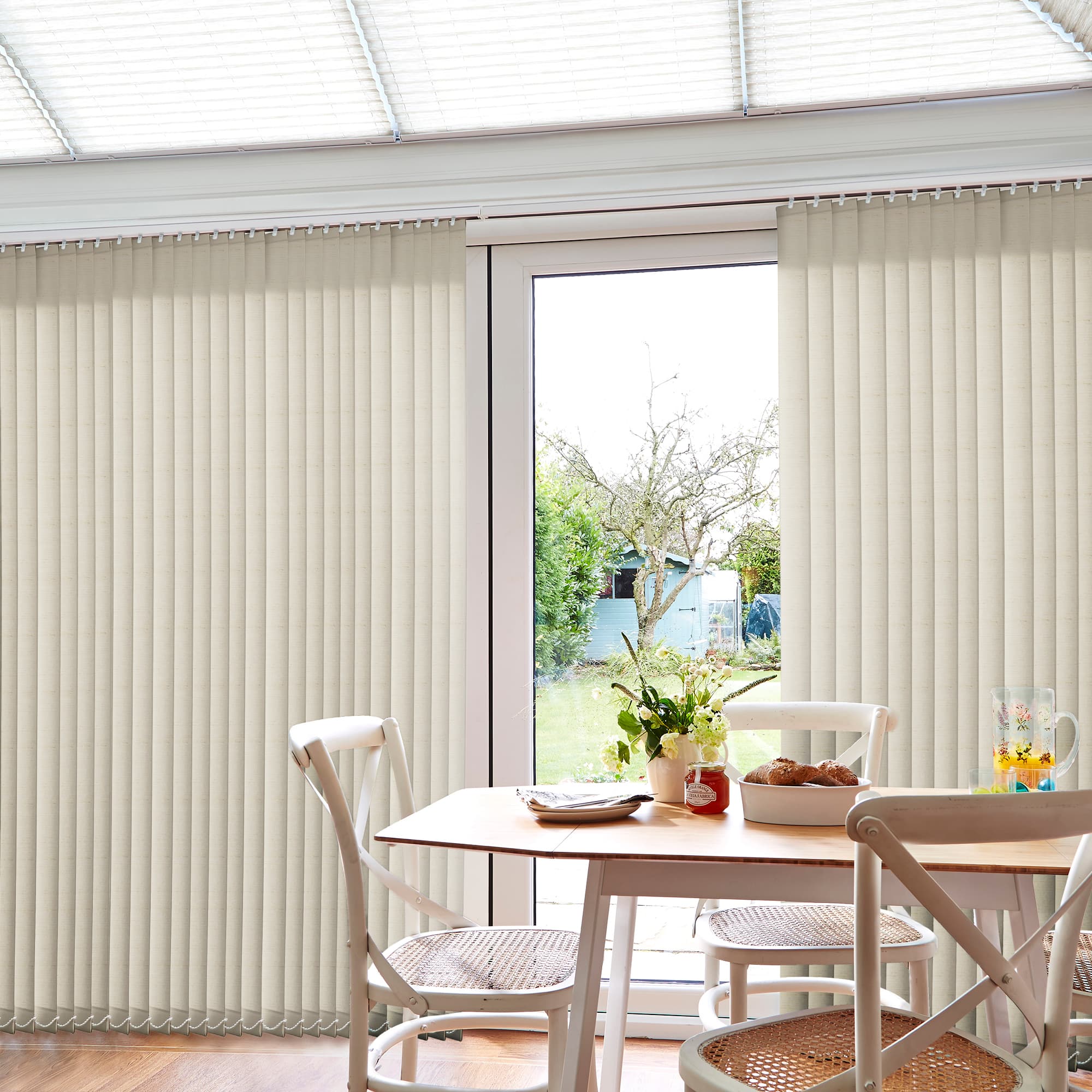 Beige vertical blinds cover sliding glass doors, panels mostly closed with a central gap, filtering daylight; small dining table and chairs nearby, sunlit conservatory/dining area overlooking a green backyard.
