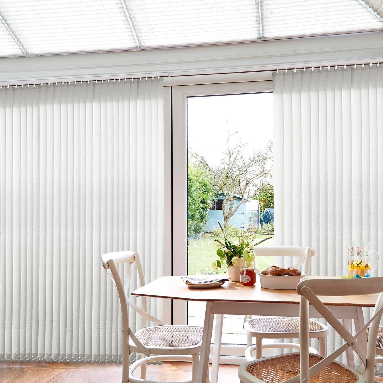 White fabric vertical blinds, floor-to-ceiling on a ceiling-mounted track, mostly closed with a central gap revealing patio door; filtering bright daylight into a sunlit dining area with table and chairs.
