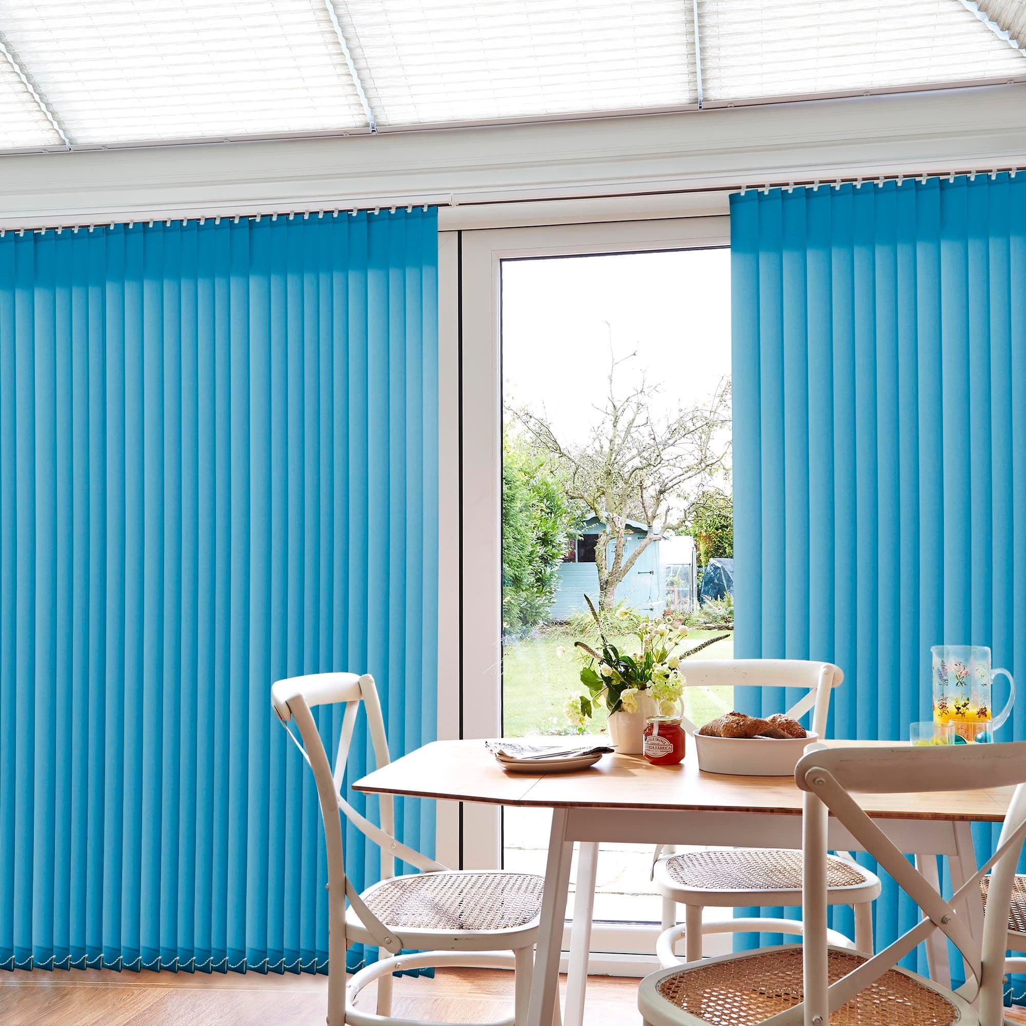 Blue vertical blinds on a track over sliding patio doors are mostly closed with a central gap letting light through; a dining table with chairs, flowers and a garden view.