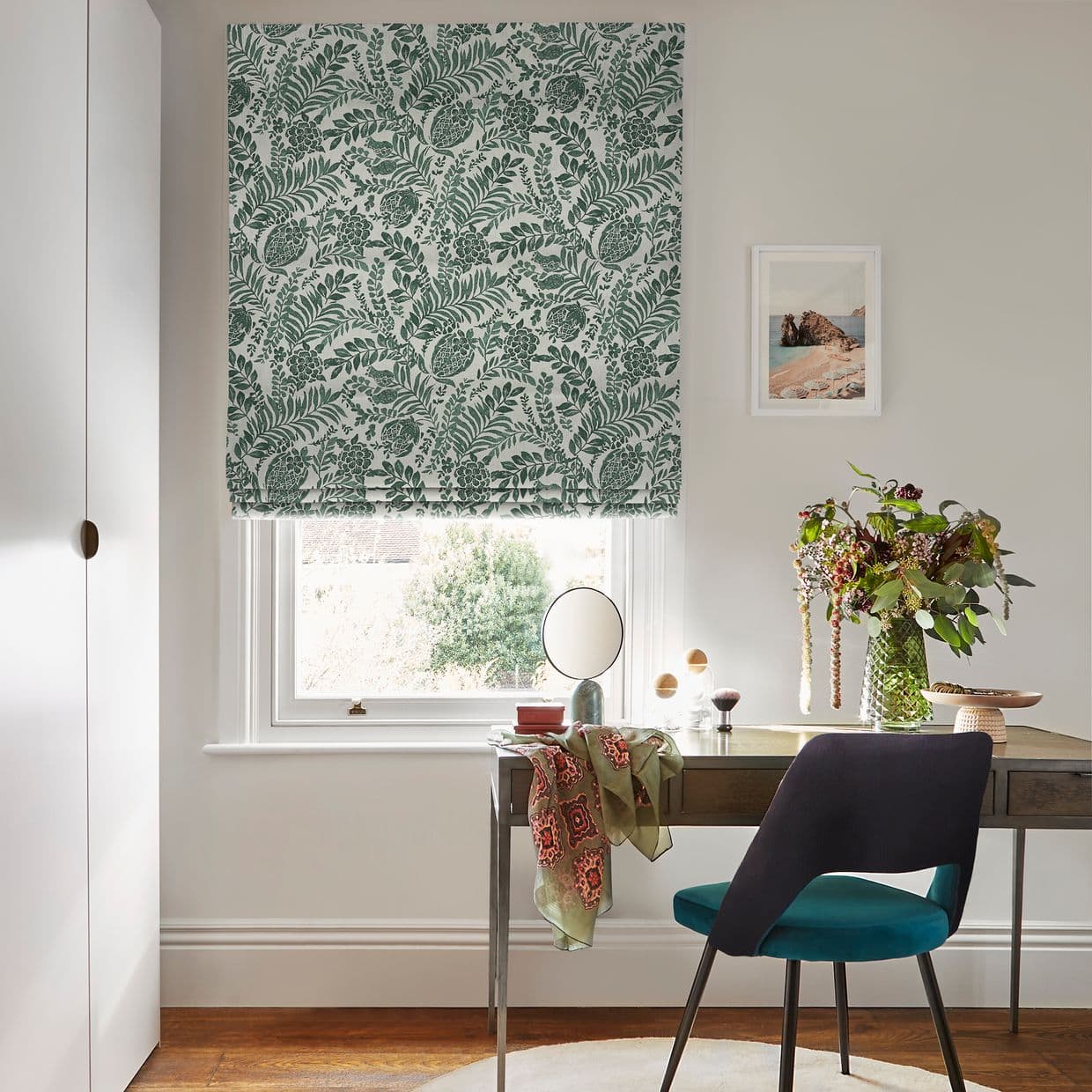 Green botanical Roman blind, lowered halfway with leafy print, filtering daylight through the window; in a bright home office with a desk, teal chair, vase of flowers and framed seascape.