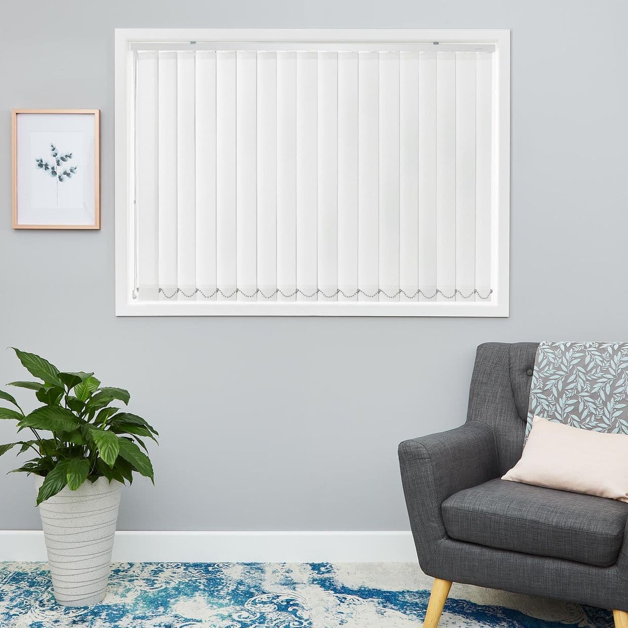 White vertical blinds covering centered window, evenly closed with bead chain pull and scalloped bottom weights; filtering soft daylight over a light-gray living room wall with armchair, plant, framed print.