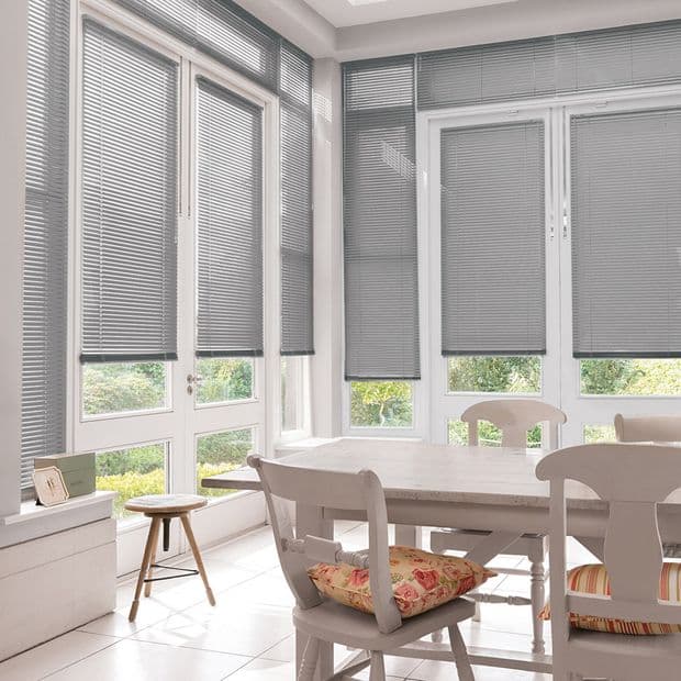 Grey horizontal Venetian blinds across tall white-framed windows and patio doors, slats mostly closed, filtering soft daylight into a sunlit dining nook with white table, chairs, tiled floor, garden glimpses.