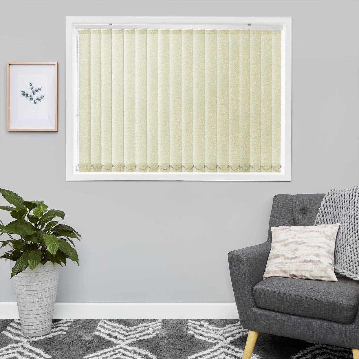 Pale textured vertical blinds cover a central window, slats closed and filtering soft daylight; neutral living room with gray walls, potted plant, framed botanical print, gray armchair with pillow.