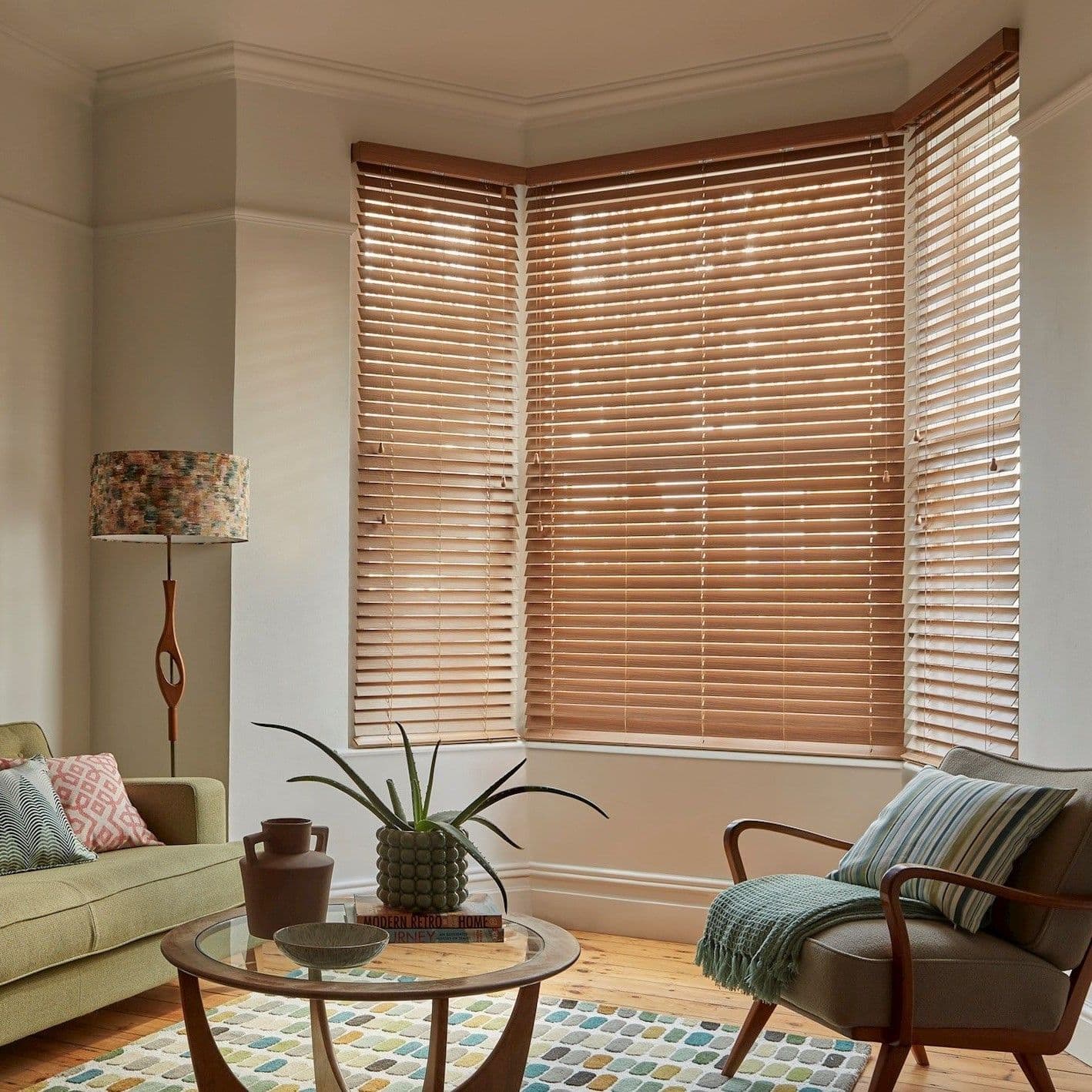Wooden Venetian blinds covering a three-pane bay window, slats mostly closed and softly filtering daylight; cozy living room with mid-century armchair, sofa, patterned rug, glass coffee table and lamp.