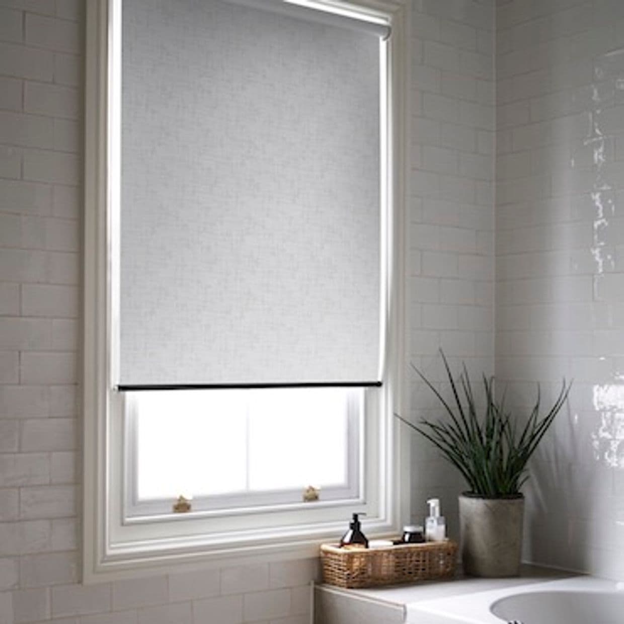 Grey fabric roller blind, lowered and covering most of the window with slight light seep at bottom; filtering daylight in a tiled bathroom above bathtub, with a potted plant and basket.