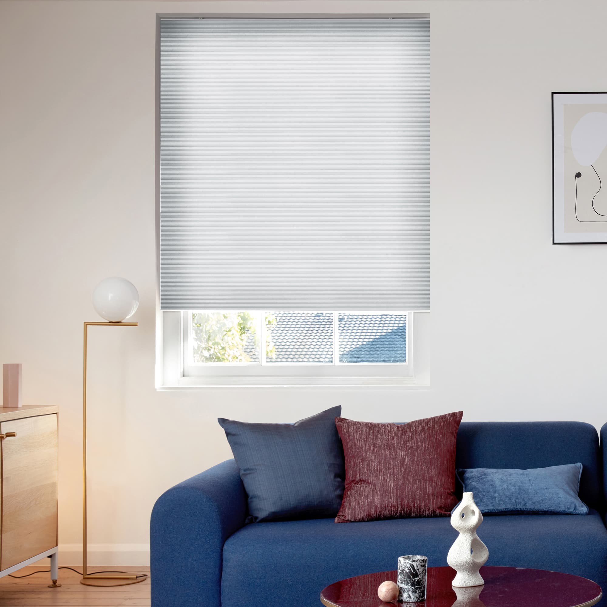 Light grey pleated cellular blind, lowered with a narrow bottom gap, filtering daylight; above a blue sofa with cushions, beside a brass floor lamp and sideboard in a living room.