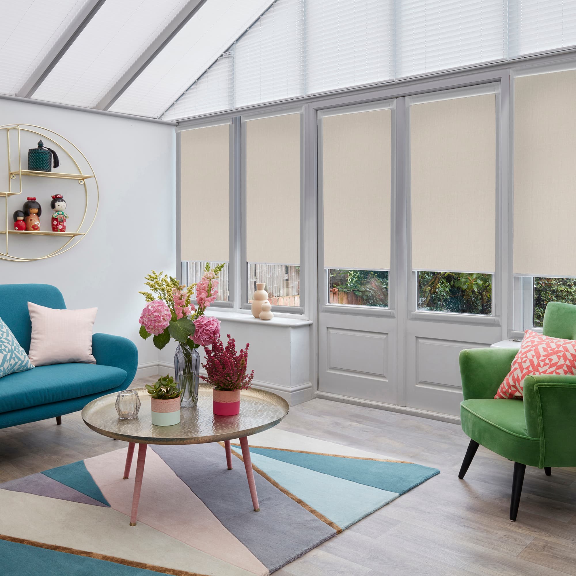 Cream roller blinds on four tall window panes, lowered to mid-height and softly filtering daylight; in a bright conservatory-style living room with skylight blinds, teal sofa and green armchair.