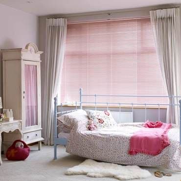 White floor-length curtains framing closed pale pink horizontal blinds, filtering soft daylight; a metal-framed daybed with floral bedding and pink throw near a wardrobe in a pastel bedroom.