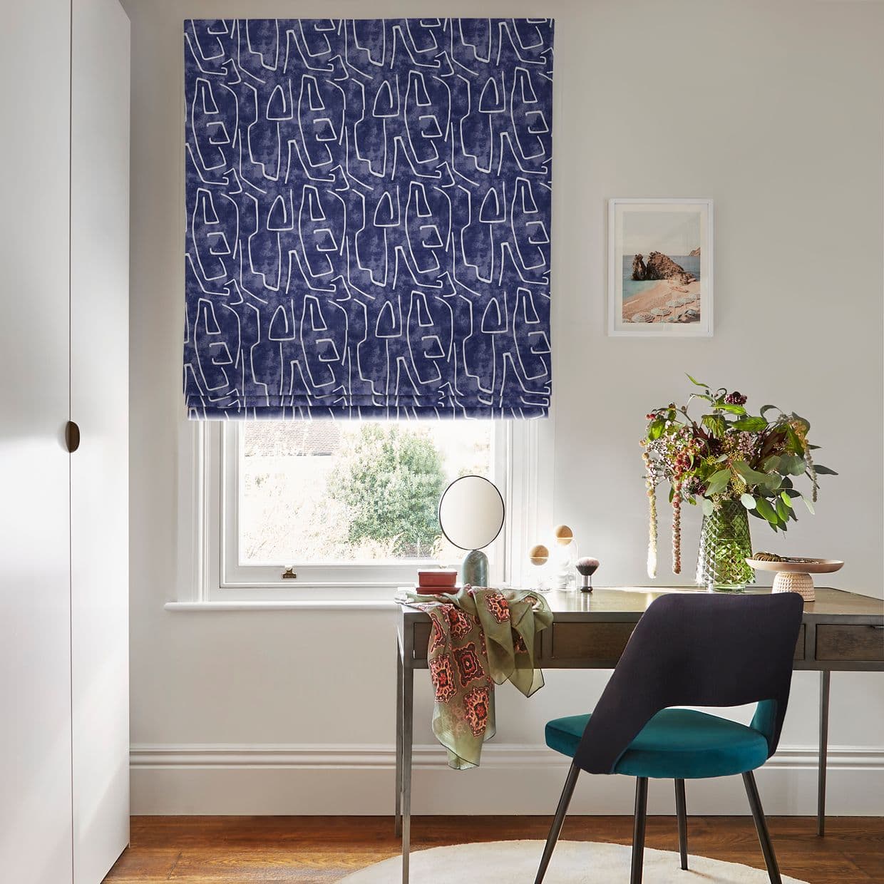 Patterned navy Roman blind covering the window, with white abstract looped motifs; lowered, filtering daylight; in a bright office with wooden desk, teal chair, vase of flowers and seaside print.