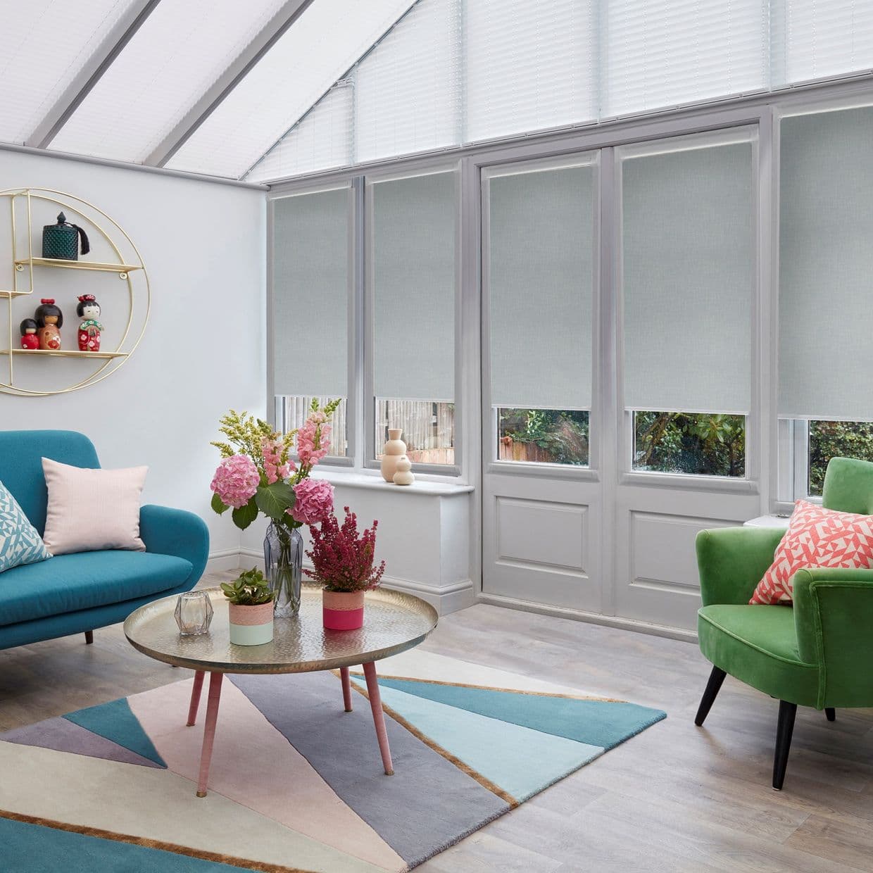 Light-gray roller blinds across tall glazed doors and roof windows, lowered mid-height filtering daylight; in a sunroom with teal sofa, green armchair, round coffee table, rug, flowers and garden glimpses.