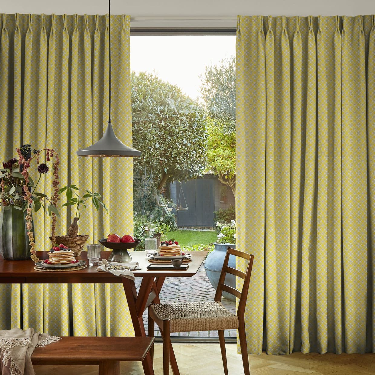 Full-length pleated yellow patterned curtains, parted centrally to reveal a glass door; filtering soft daylight. Dining table set with pancakes and fruit, pendant lamp overhead, view of a green backyard.