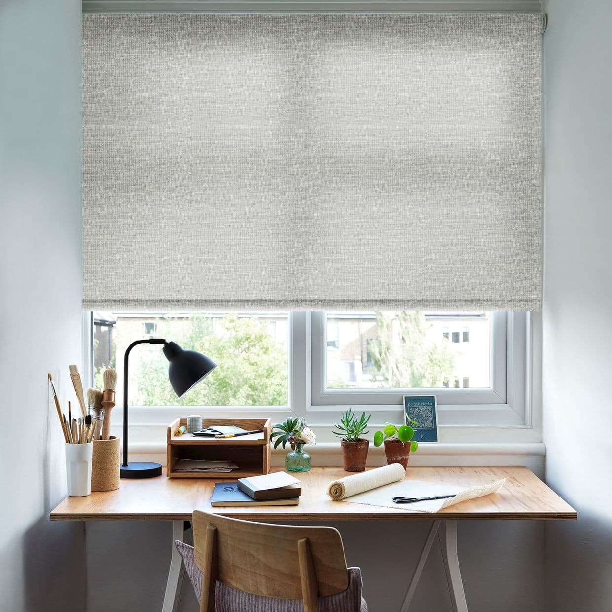 Light-grey textured roller blind lowered almost to the sill, its woven fabric filtering soft daylight; a home workspace sits beneath â€” wooden desk with lamp, brushes, notebooks and potted succulents.