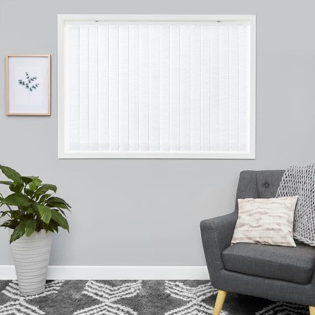 White vertical blinds inside a white-framed window, closed and filtering soft daylight; set in a grey-walled living room with a potted plant, framed botanical print, grey armchair and patterned rug.