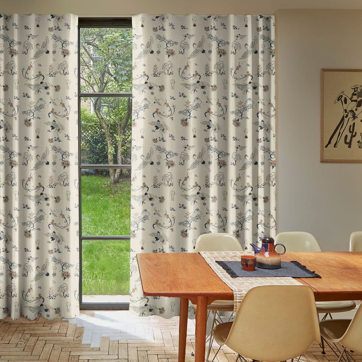 Cream printed curtains (floral and bird pattern) drawn aside over a tall black-framed window, filtering daylight and revealing a grassy backyard; dining table and chairs in a warm, softly lit room.