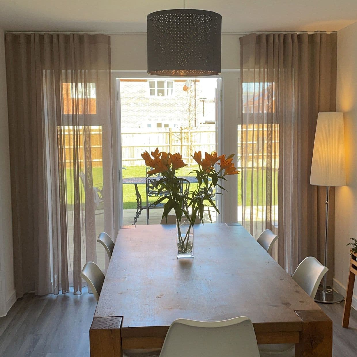 Sheer beige, floor-to-ceiling pleated curtains drawn across sliding glass doors, softly filtering daylight; dining room with a large wooden table, vase of orange lilies, white chairs and backyard lawn beyond.