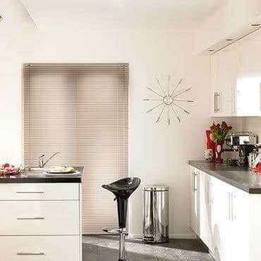 Beige horizontal venetian blind covering window, closed and filtering soft daylight; modern white kitchen with black countertop, bar stool, stainless bin, starburst wall clock and vase of red tulips.