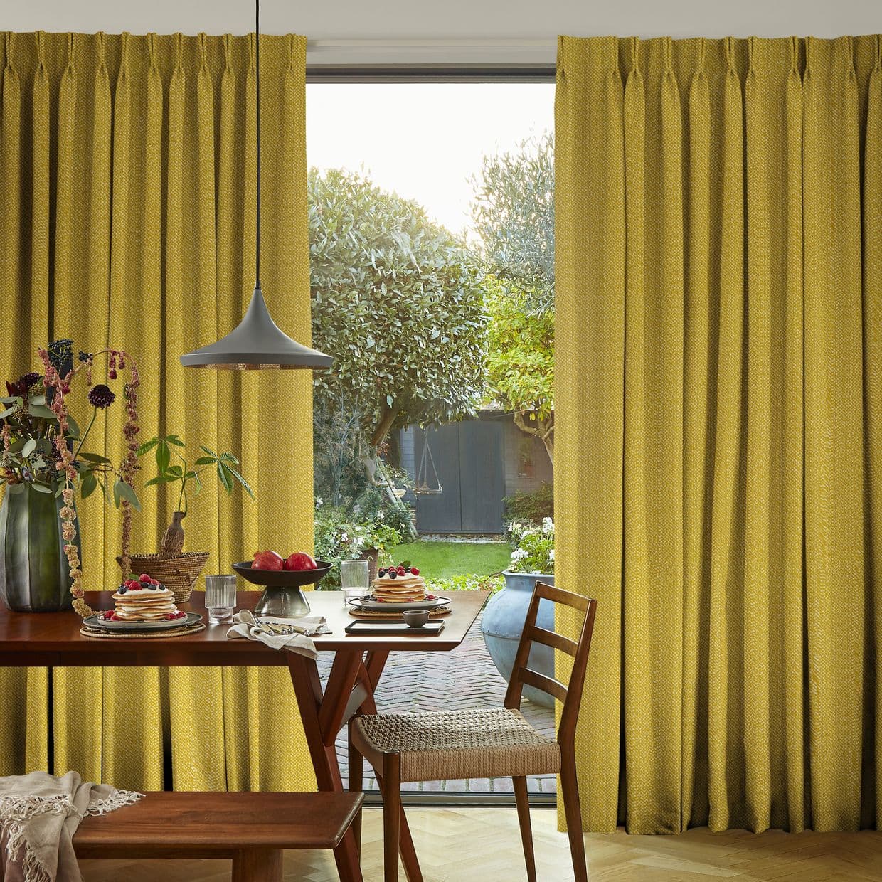 Floor-length mustard-yellow pleated curtains, drawn aside in gathered folds, frame a glass sliding door, admitting soft daylight onto a dining table with pancakes and a lush backyard garden.