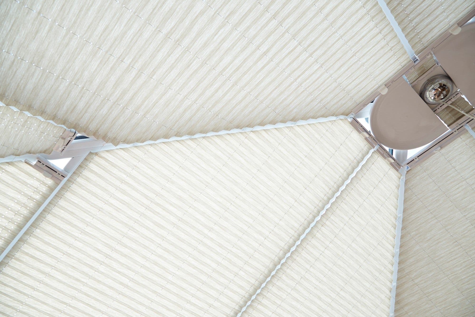 Beige pleated cellular blinds covering triangular skylight panels, attached to white frame and rails; closed and diffusing soft daylight across a ceiling-mounted metal skylight structure.