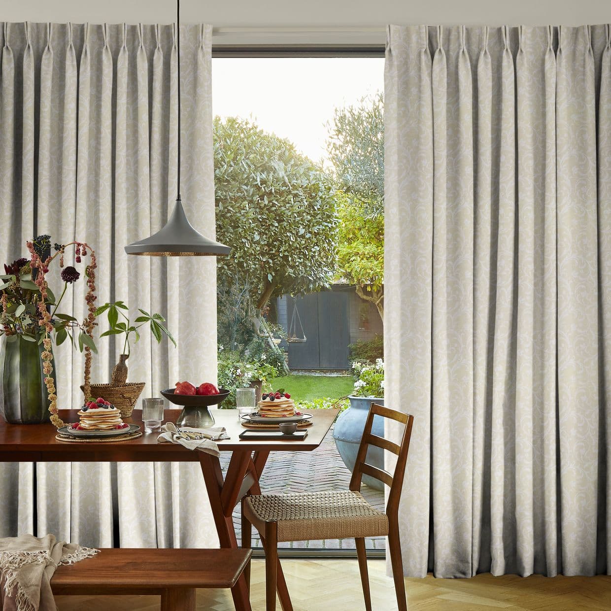 Pleated cream patterned curtains pulled aside, framing a sliding door and admitting daylight; a wooden dining table set for breakfast under a pendant lamp, opening onto a leafy backyard.