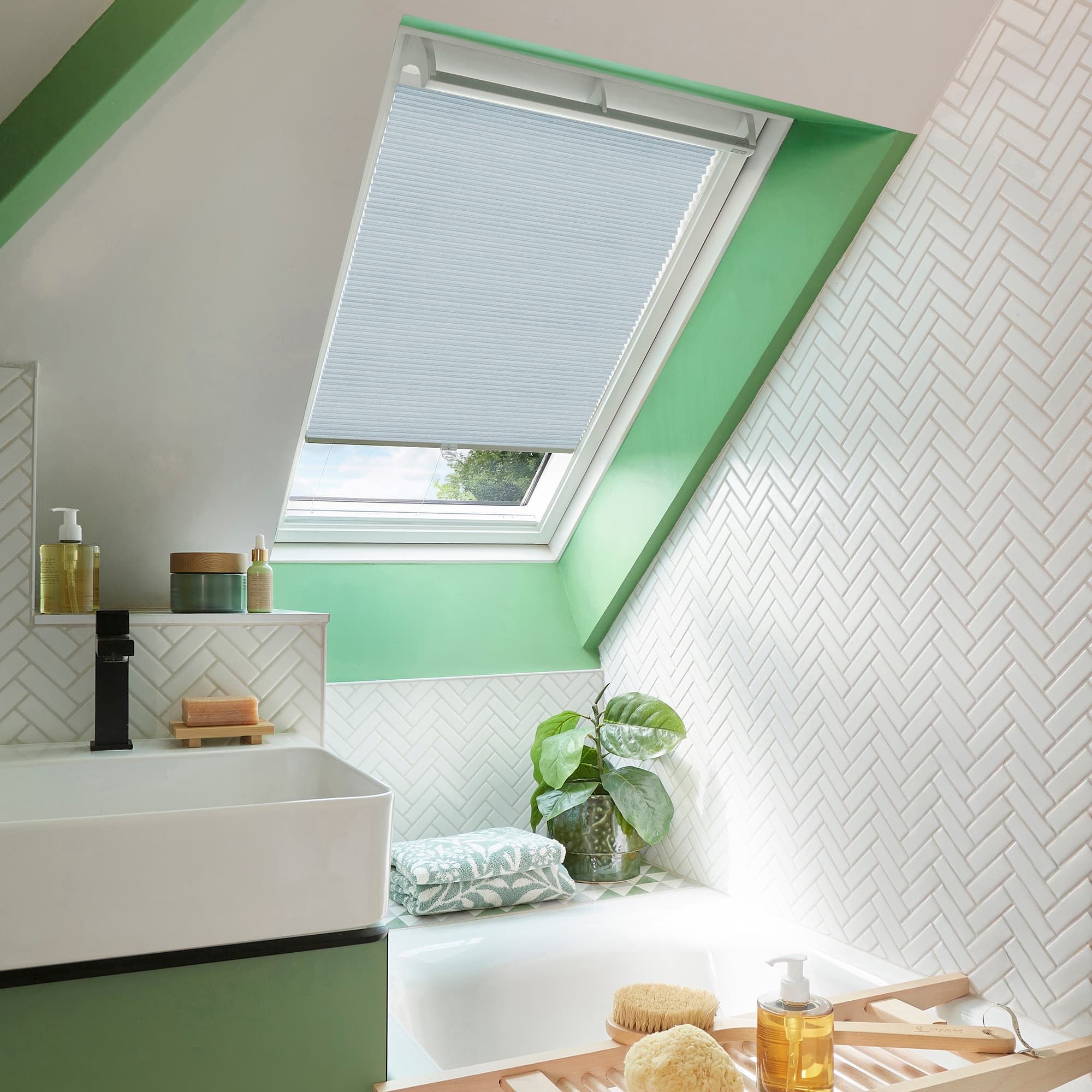 A white pleated skylight blind, lowered and filtering daylight, covers a sloped window in a bright attic bathroom with green trim, white herringbone tiles, sink, bath, plant and toiletries.