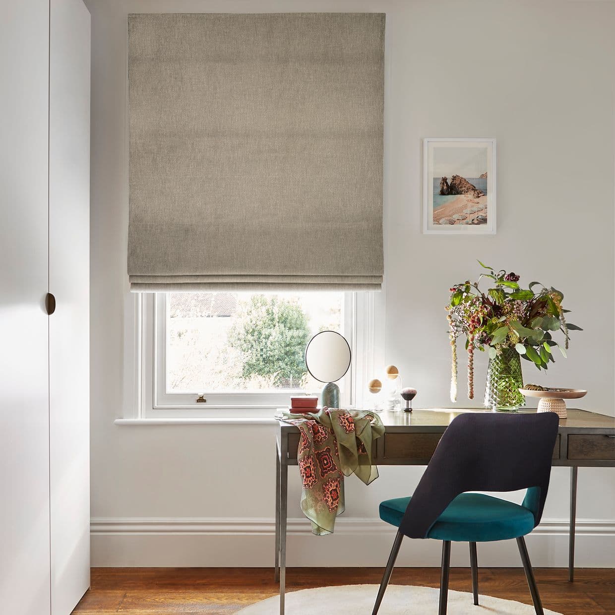 Beige linen Roman blind, lowered in soft horizontal folds, filtering bright daylight through a lower gap; above a desk in a light-filled home office with a teal chair, vase of flowers.