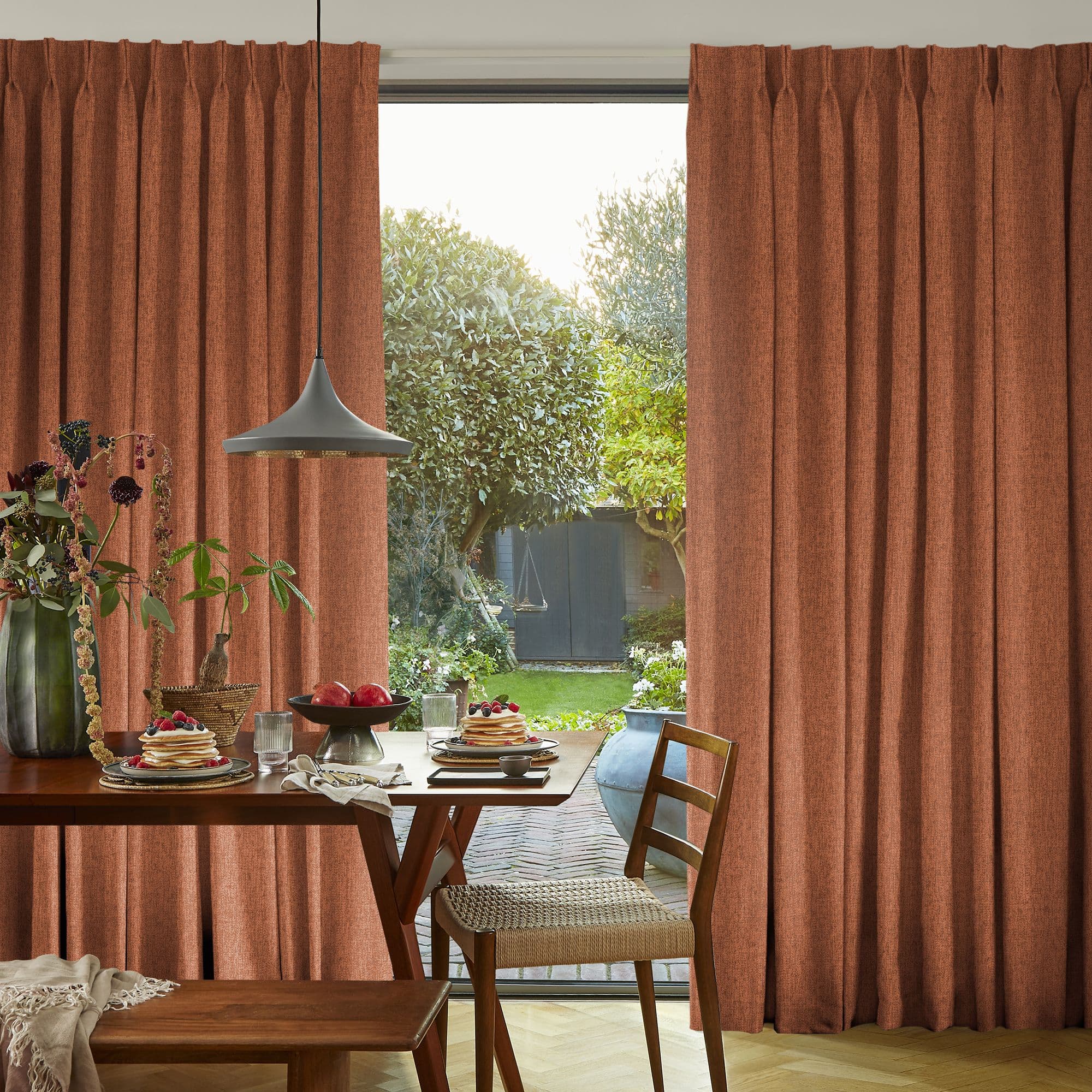 Rust-colored pleated, floor-length curtains, parted at center to reveal sliding glass door; curtains filter warm light. Wooden dining table set for breakfast faces a sunlit backyard garden.